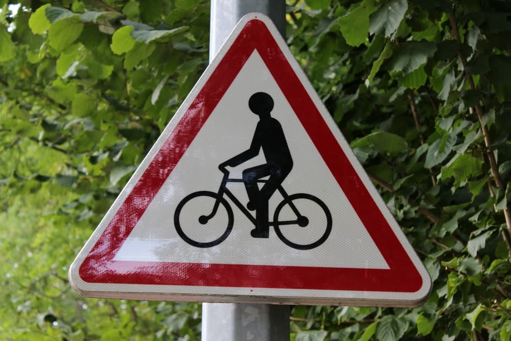 A triangular road sign with a bicycle symbol, surrounded by green foliage in the background.