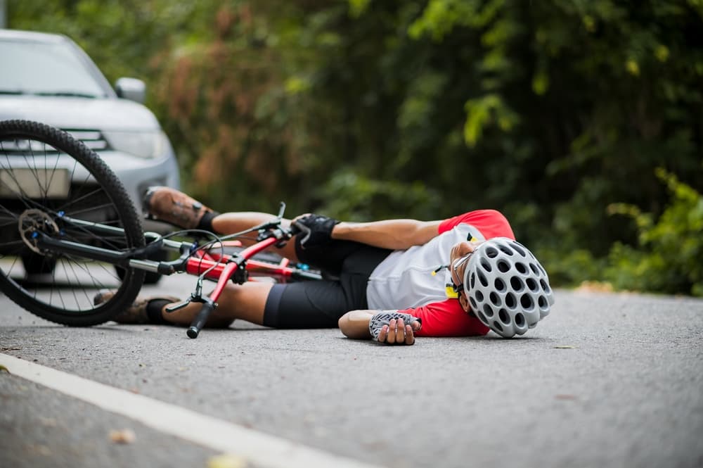 A cyclist lying injured on the road beside a fallen bicycle, with a car nearby.