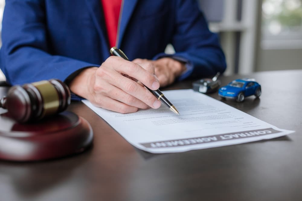 Person signing a contract at a desk with a gavel, toy cars, and car keys beside them.