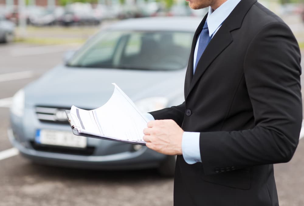 Man in a suit reviewing documents while standing in front of a car in a parking lot.