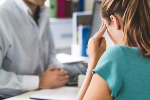 A woman in a doctor's office, holding her head in discomfort, as she discusses her symptoms with a healthcare professional