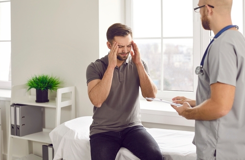 A man sitting on a medical examination table, holding his head in pain while talking to a doctor in a bright, white room.