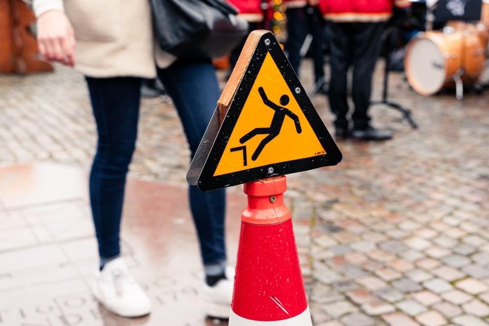 A yellow slippery caution sign is on the ground. The sign is placed on a sidewalk